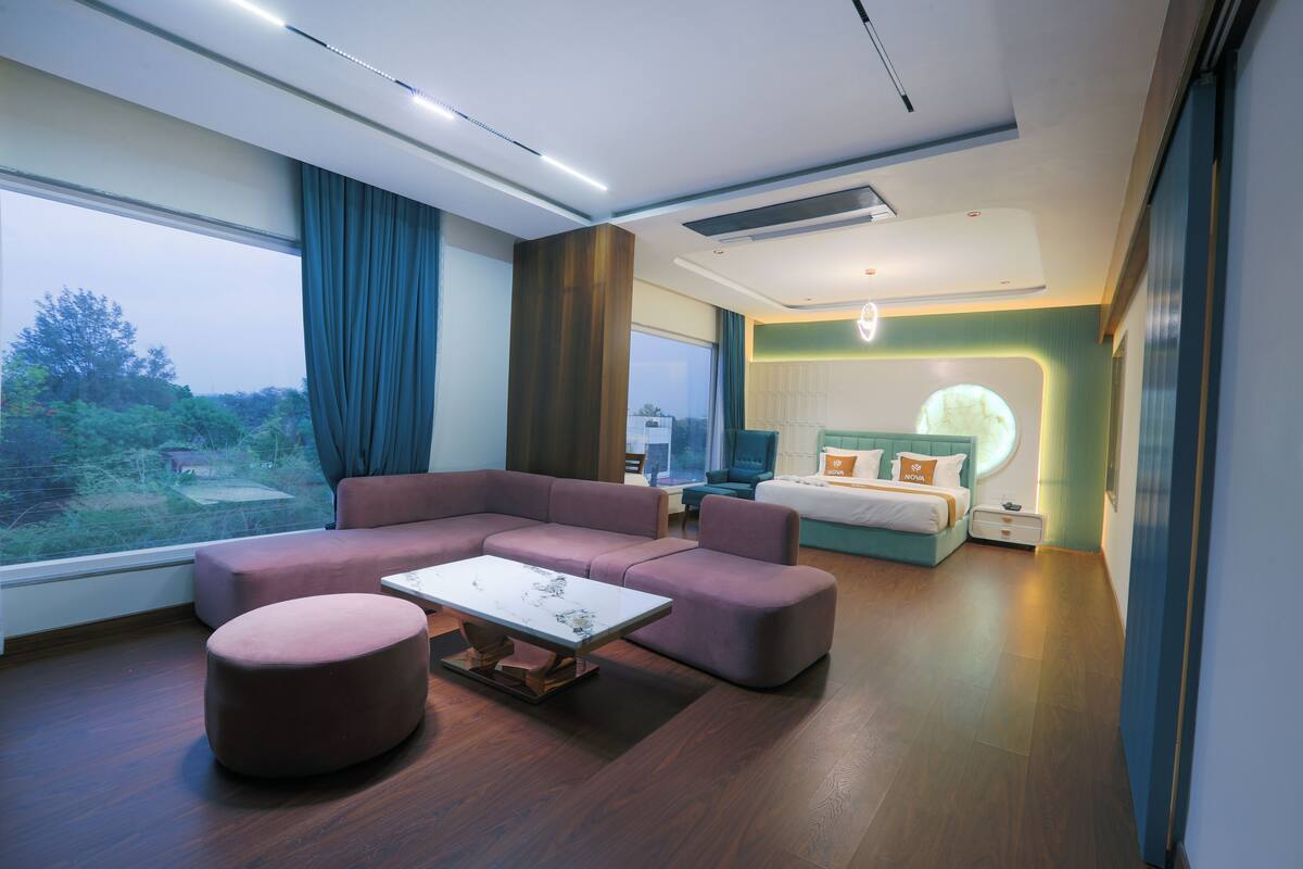 Una habitación de hotel moderna con un sofá modular, un reposapiés redondo, una mesita, una cama con cabecera y un ventanal con vistas al exterior.