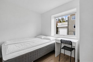 A bedroom with a bed, a desk, and a chair, a window with a view of buildings, and a white wall.