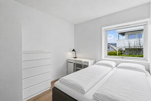 Um quarto com uma cama, uma escrivaninha, uma cadeira, um abajur e uma janela com vista para edifícios.