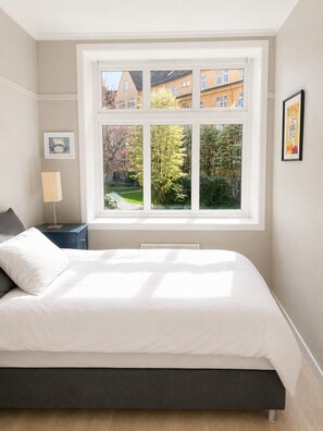 Ein Schlafzimmer mit großem Fenster, einem Bett, einem Nachttisch, einer Lampe und einem gerahmten Bild an der Wand.