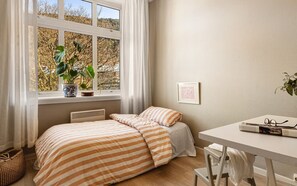 Ein Schlafzimmer mit Bett, einem Fenster mit Vorhängen, einem Nachttisch mit Gläsern und einem gerahmten Bild an der Wand.