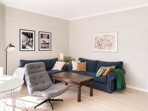 Ein Wohnzimmer mit einem blauen Sofa, einem grauen Sessel, einem Holztisch und einem weißen Tisch. An der Wand hängen zwei gerahmte Bilder, eine Stehlampe und eine grüne Decke liegt auf dem Sofa.