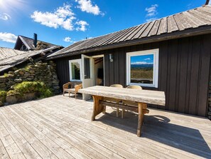 A wooden deck with a table and chairs, a stone wall, and a view of mountains through a window.