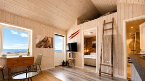 Ein Zimmer mit Holzwänden, einem Fernseher, einem Schreibtisch, einem Stuhl, einem Bett und Blick ins Freie.