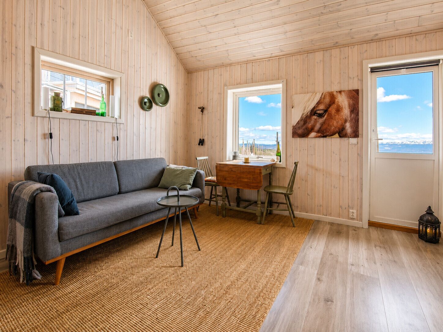 Une pièce chaleureuse avec un plafond en bois, un canapé gris, une petite table ronde, une table à manger en bois avec des chaises, et une grande fenêtre offrant une vue sur le ciel et les montagnes.