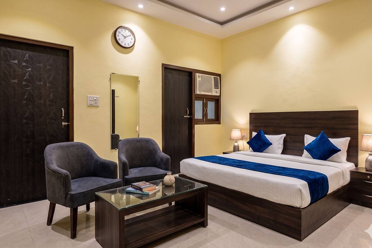 Habitación de hotel con una cama, dos sillones, una mesa de centro de vidrio, una mesita de noche con lámpara, un reloj de pared y un aire acondicionado.