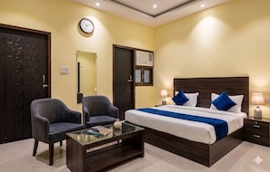 A hotel room with a bed, two armchairs, a glass coffee table, a bedside table with a lamp, a wall clock, and an air conditioning unit. - Shri Amar Dham Near Prem Mandir Vrindavan (Vrindavan)
