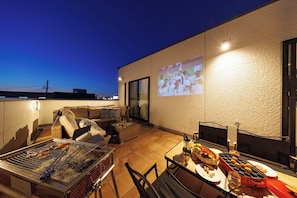 Eine Terrasse im Freien mit Grill, Sitzgelegenheit und einer Leinwand, die ein Sportevent zeigt.