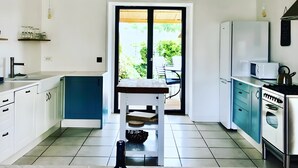 Una cocina con armarios blancos, encimera azul, lámpara colgante de mimbre y una botella de vino sobre la mesa.