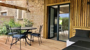 Una terraza de madera con una mesa redonda y sillas, un muro de piedra y un ventanal corredero que da a un espacio al aire libre.