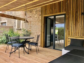Una terraza de madera con una mesa redonda y sillas, un muro de piedra y un ventanal corredero que da a un espacio al aire libre.