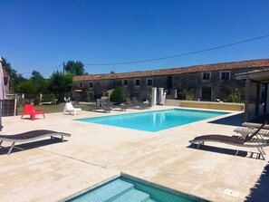 Una piscina con sillas de descanso, una silla roja y un edificio de piedra con techo de tejas al fondo.