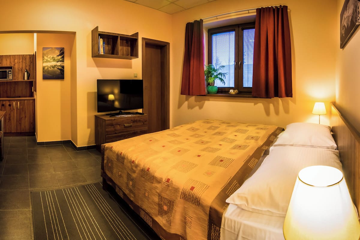 Una habitación de hotel con cama, televisión, ventana con cortinas, lámpara y un pequeño estante con libros.