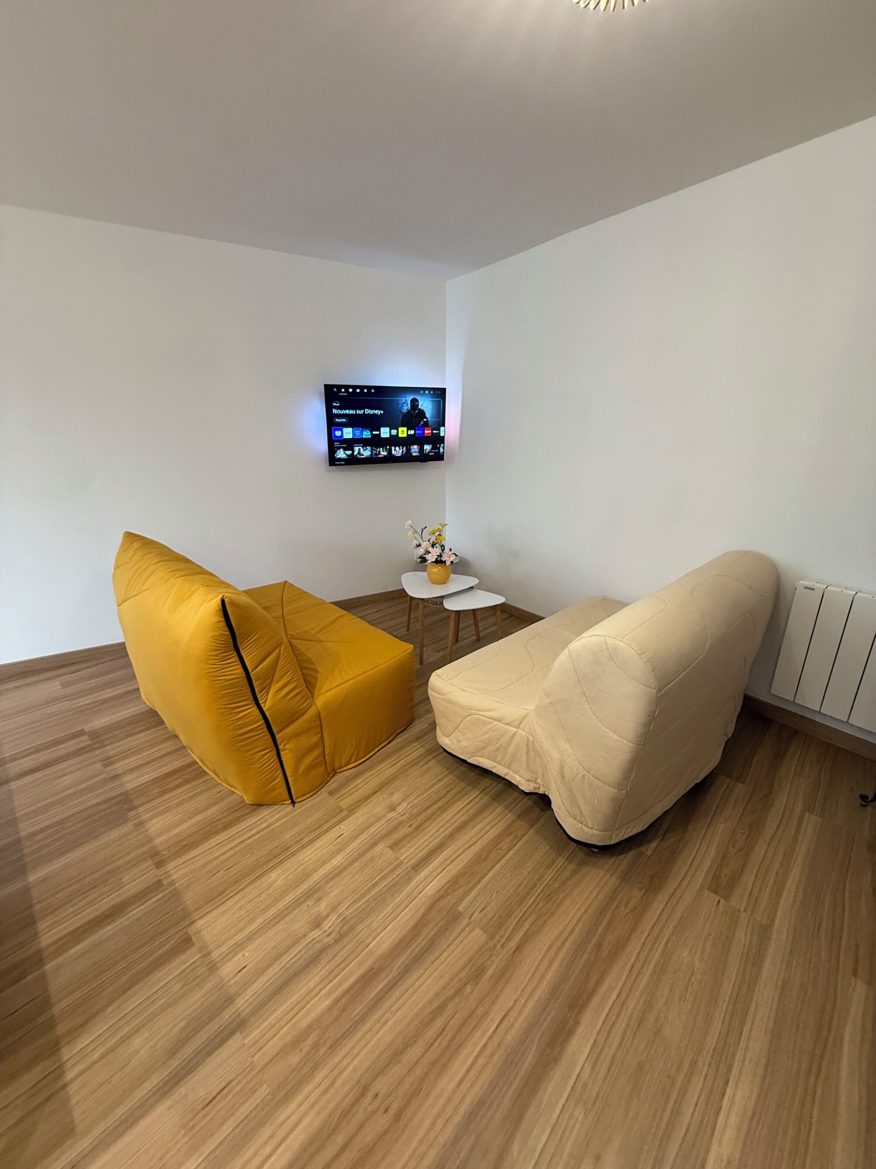 Una sala moderna con un televisor de pantalla plana montado en la pared, una pequeña mesa blanca con un florero y dos sofás, uno amarillo y otro color beige.