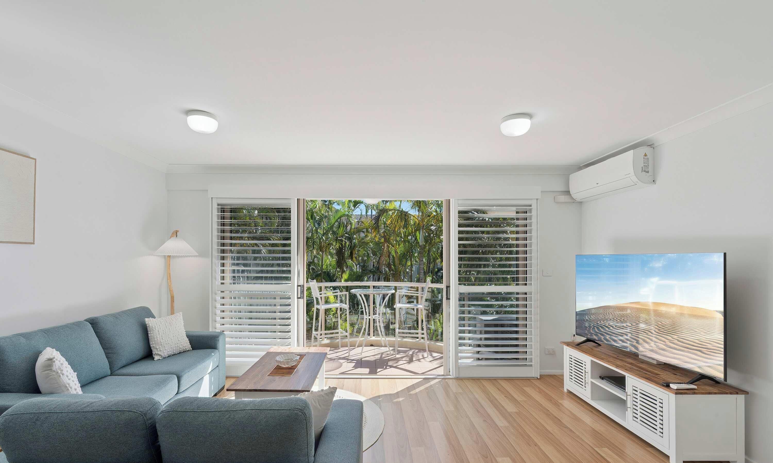 A modern living room with a sofa, TV, and a balcony with white shutters.