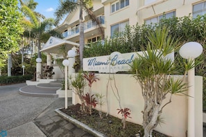Ein Hotel Eingang mit einem Schild, auf dem 'Miami Beach Holiday Apartments' steht, einer geschwungenen Auffahrt und einer begrünten Anlage mit verschiedenen Pflanzen.