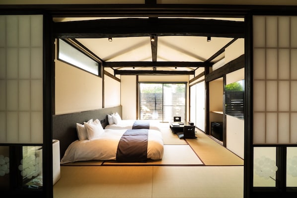 Ein traditioneller japanischer Raum mit Tatami-Boden, einem Bett mit weißer und grauer Bettwäsche, einem kleinen Schreibtisch mit Stuhl und einer Schiebetür zu einem Balkon.