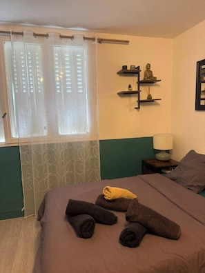 A bedroom with a bed, a window with curtains, a nightstand with a lamp, and a wall-mounted shelf with decorative items.