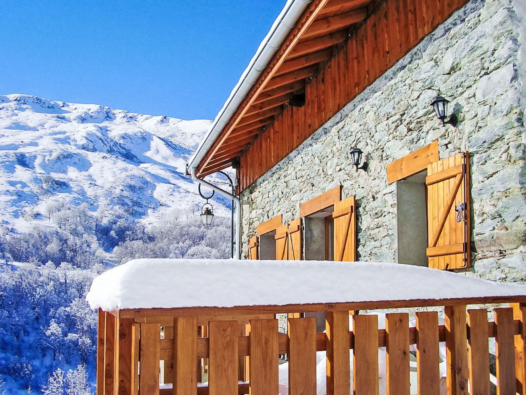 Un edificio de piedra con contraventanas de madera, un balcón cubierto de nieve y montañas al fondo.