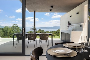 A dining area with a table set for four, a kitchen area with a sink and counter, and a view of the landscape. - Villa Serenis – a Modern New-Build with Private Pool and Breathtaking Sea Views (Labin)