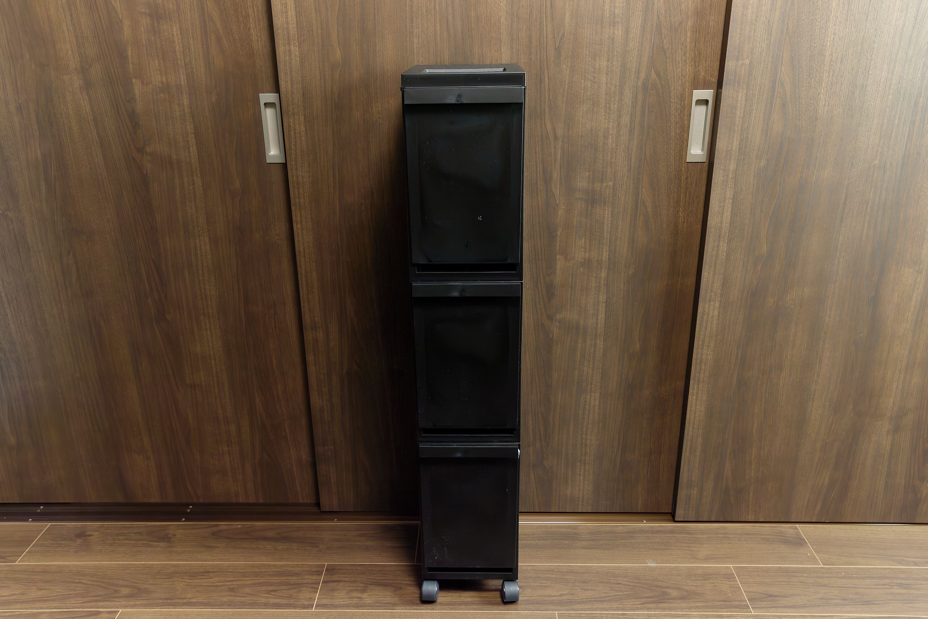A black, three-tiered, wheeled storage unit against a wooden wall.