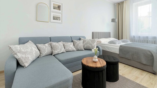 Una habitación de hotel moderna con un sofá gris, una cama, una mesita redonda de madera y una mesita de noche.