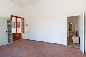 An empty room with a tiled floor, a wooden door, a white wall, and a small wooden cabinet. - GuestHost - Casa degli Oleandri (Naxos)