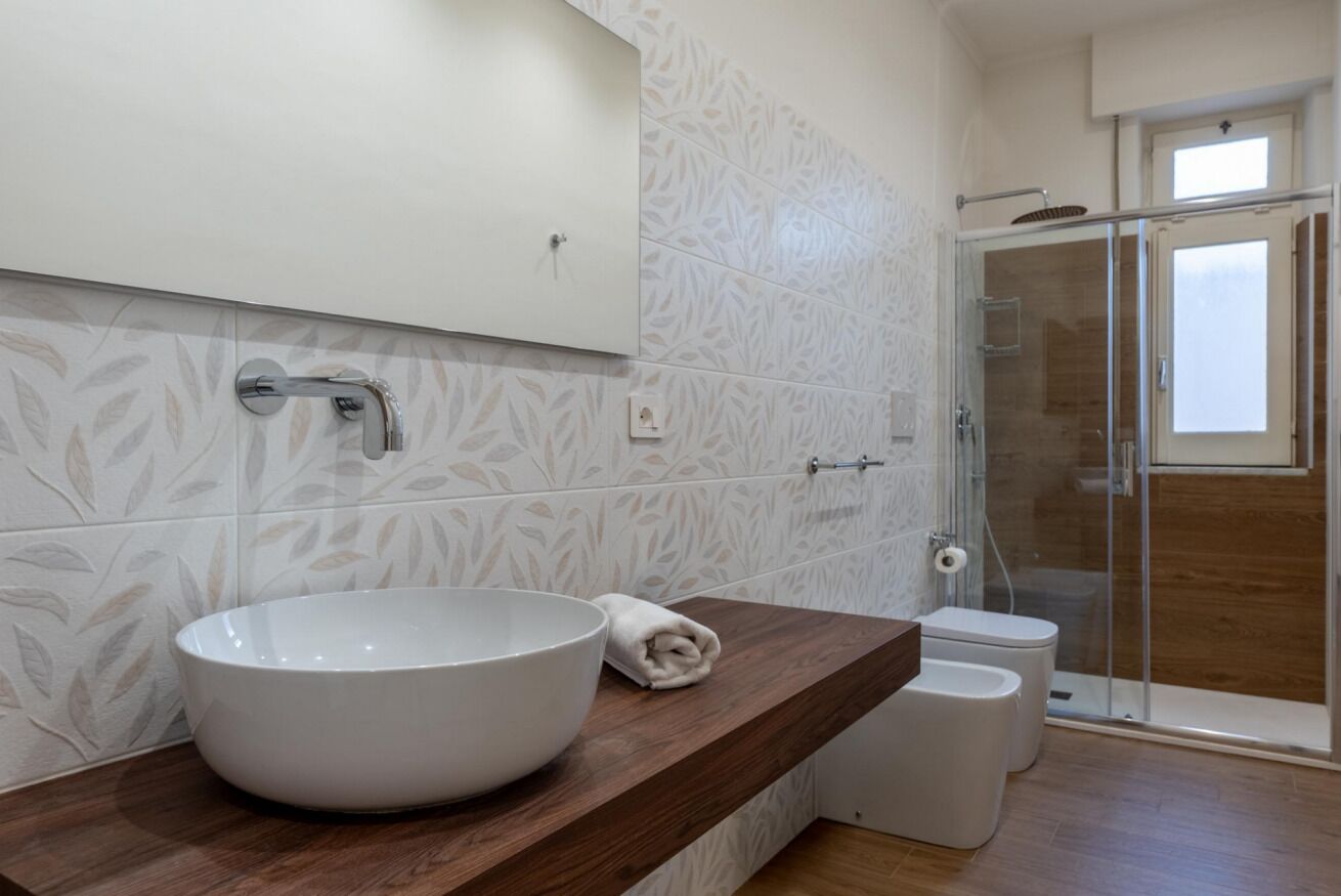 Ein Badezimmer mit einem großen weißen Waschbecken, einer Holz-Arbeitsplatte, einer Duschkabine aus Glas und Wandfliesen mit Muster.
