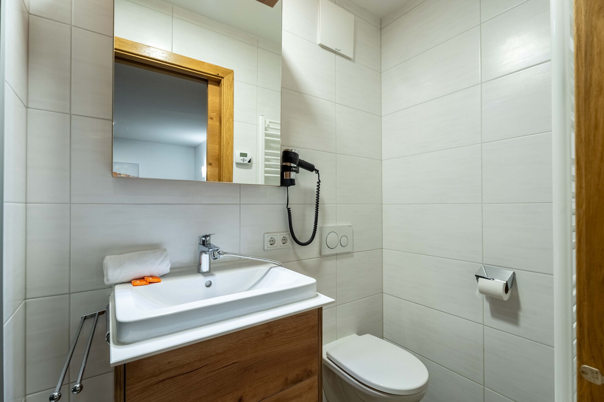 A bathroom with a white sink, wooden vanity, a mirror, a toilet, and a hairdryer mounted on the wall.