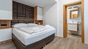 Un dormitorio moderno con una cama grande, cabecera integrada, mesitas de noche y una puerta de madera que da a un baño con espejo y lavamanos.