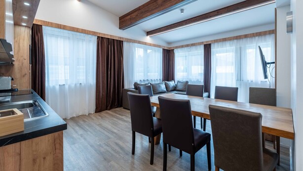 Una cocina moderna con estufa integrada, un comedor con mesa y sillas de madera, y ventanas grandes con cortinas.