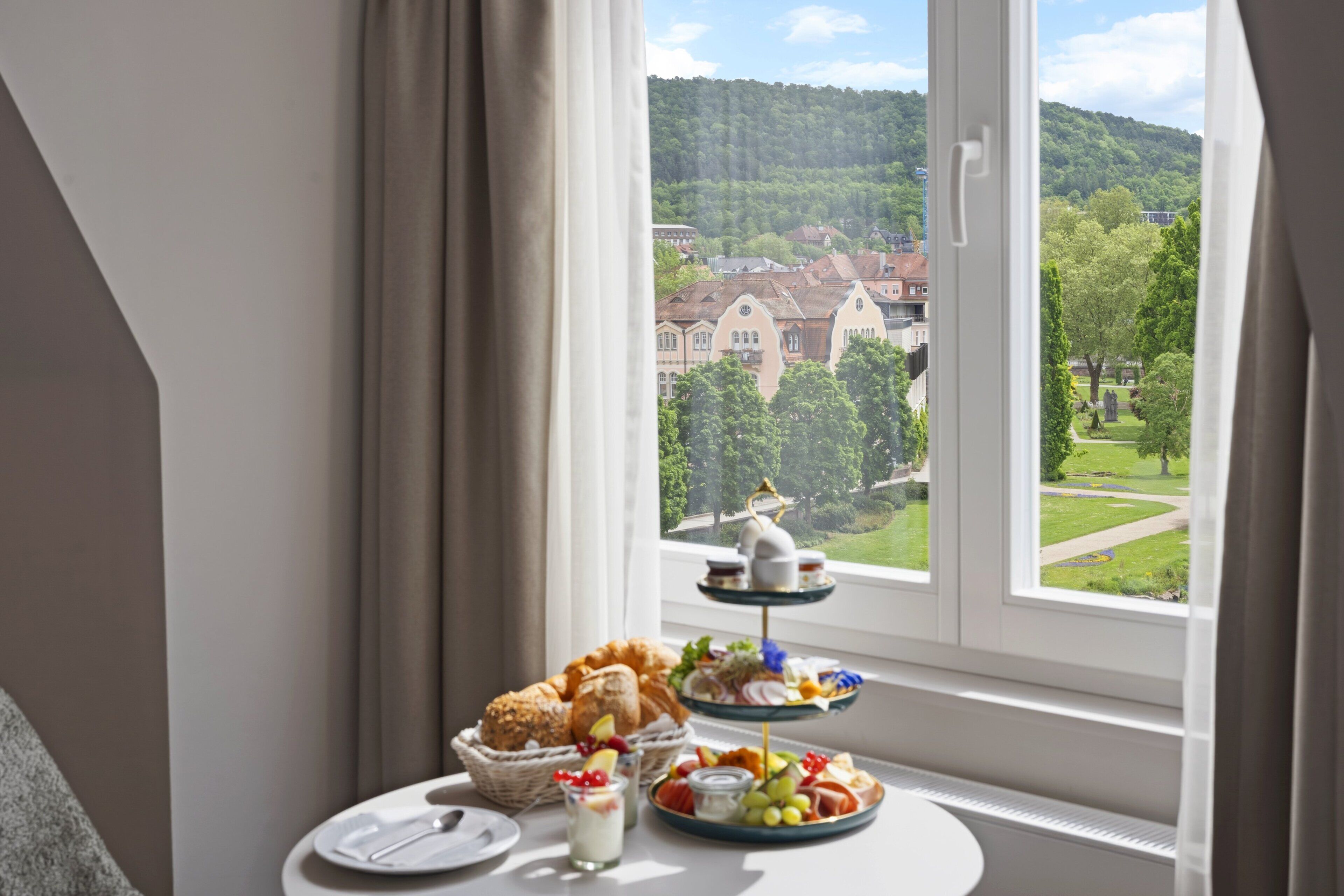 A breakfast setup with pastries, fruits, and dairy on a table by a window with a view of a town and hills.