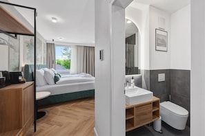 A modern hotel room with a bed, a wooden desk, a bathroom with a sink and toilet, and a view of greenery outside. - Schweizerhaus Bad Kissingen (Bad Kissingen)