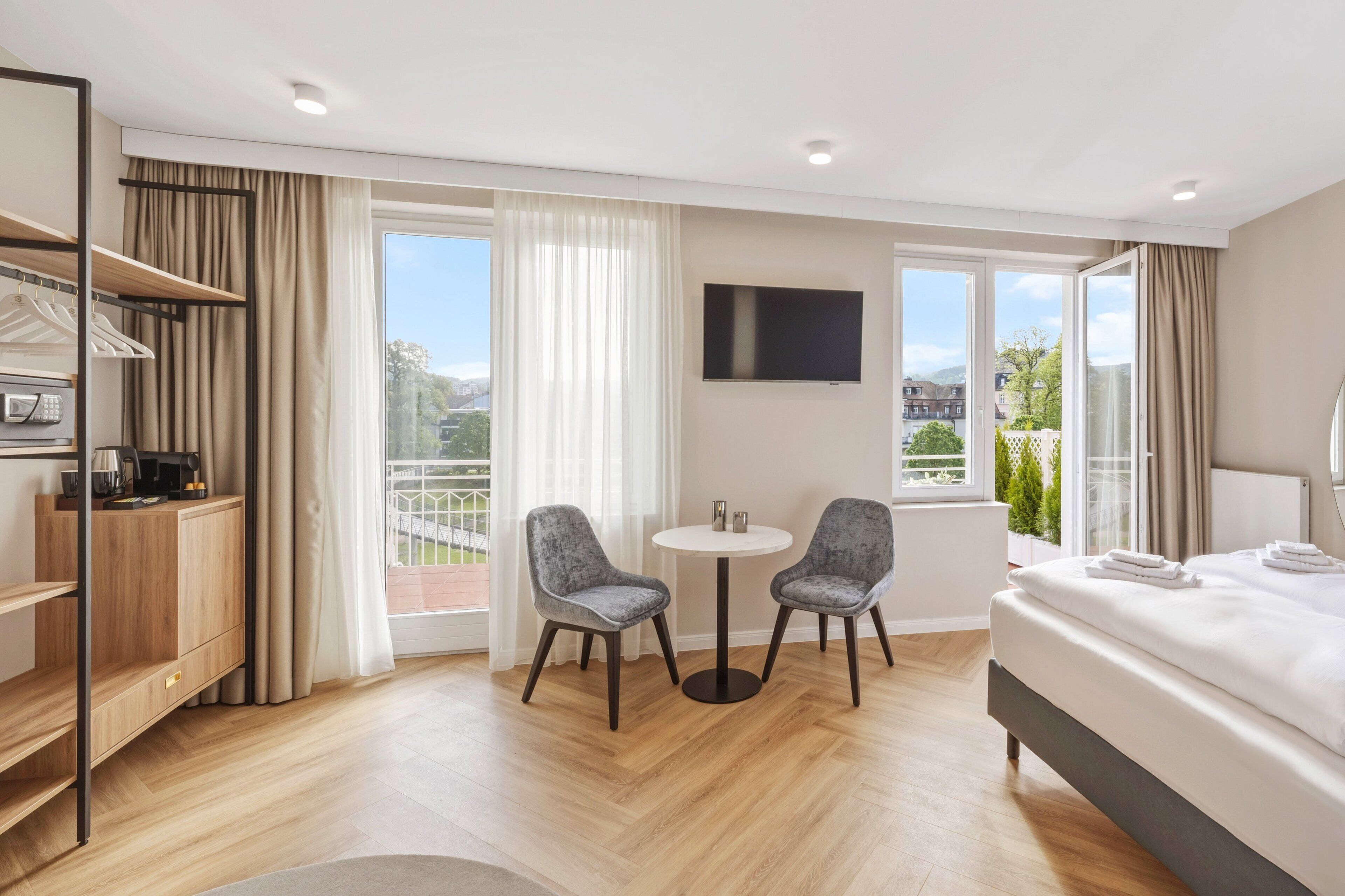 A hotel room with a bed, two chairs, a small table, a TV, and a balcony with curtains.