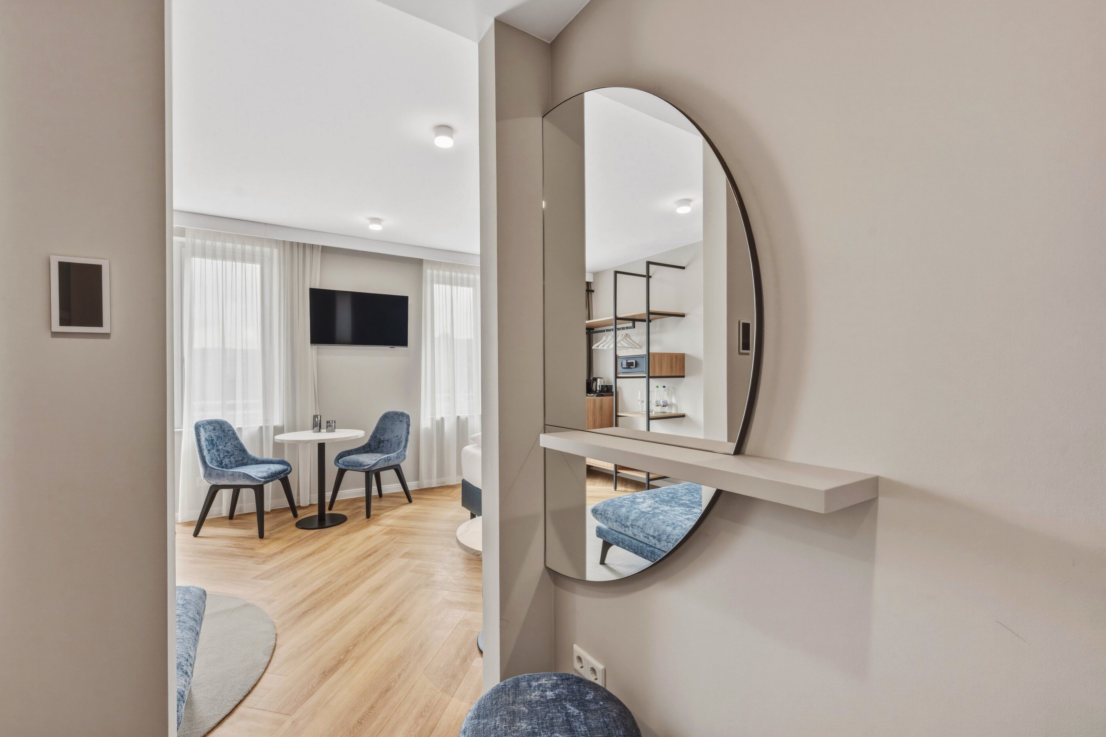 A modern hotel room with a large mirror, a flat-screen TV, a dining area with chairs, and a wooden floor.