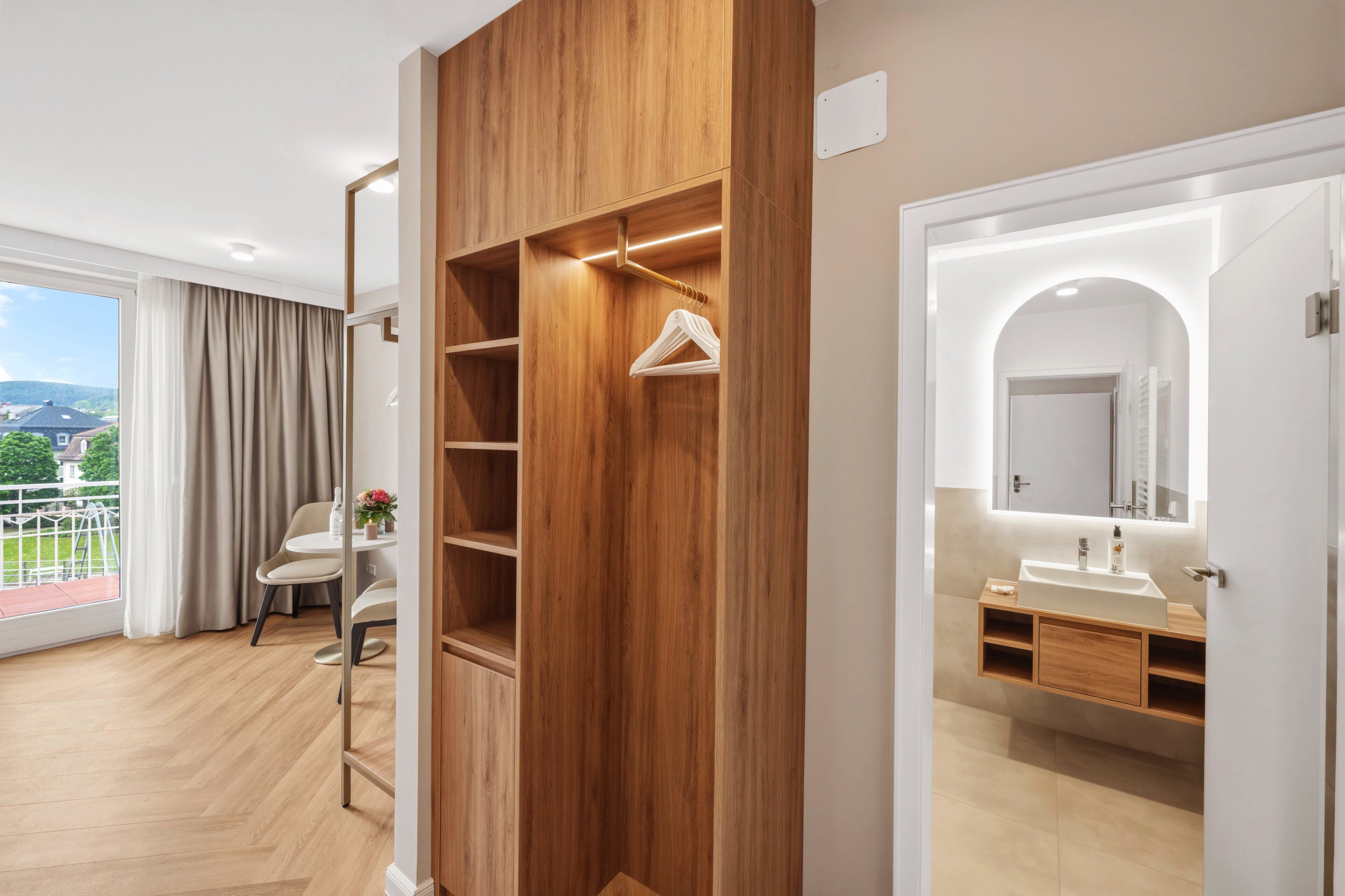 Ein modernes Hotelzimmer mit Holzschrank, einem Balkon mit Aussicht, einem Badezimmer mit Waschbecken und Spiegel sowie einem Essbereich mit Tisch und Stühlen.