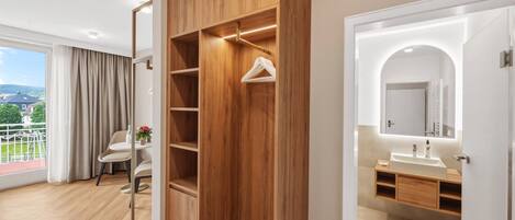 Ein modernes Hotelzimmer mit Holzschrank, einem Balkon mit Aussicht, einem Badezimmer mit Waschbecken und Spiegel sowie einem Essbereich mit Tisch und Stühlen.