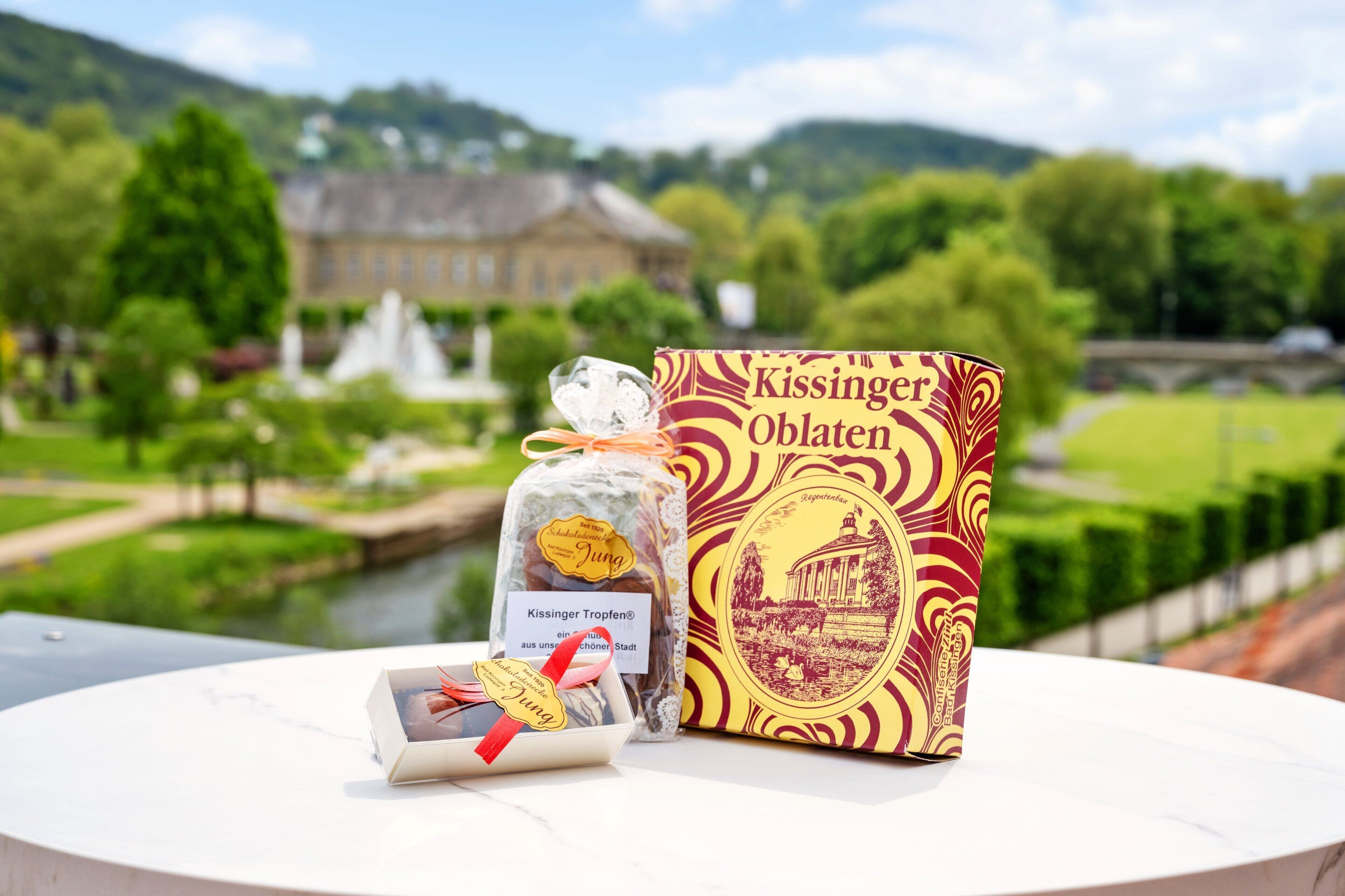 A table with a box labeled 'Kissing Oblaten' and a small gift bag with a red ribbon, set against a scenic outdoor background with a river and buildings.