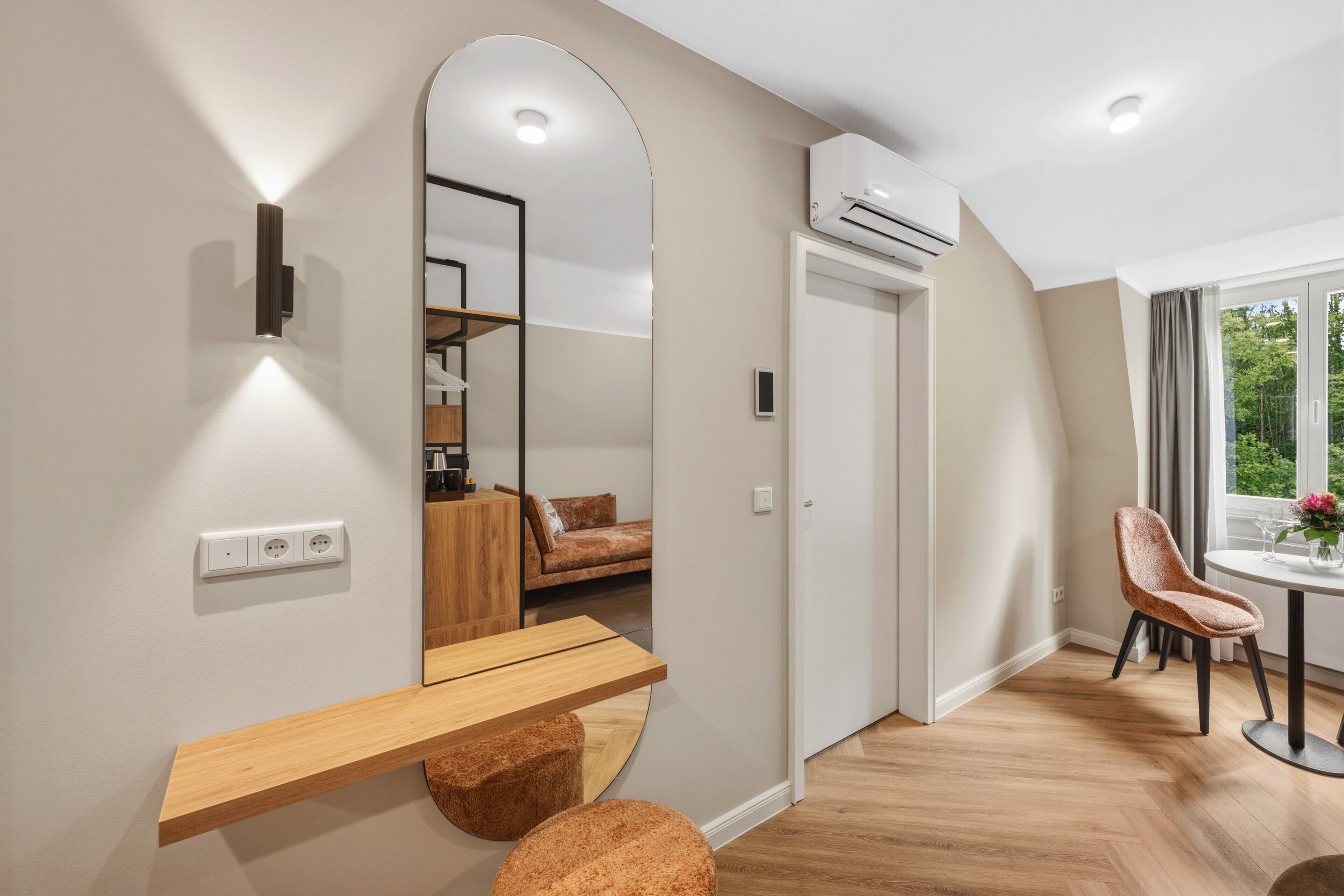 A modern room with a wooden desk, a round orange chair, a mirror, and a view of a bedroom with a bed and a window.