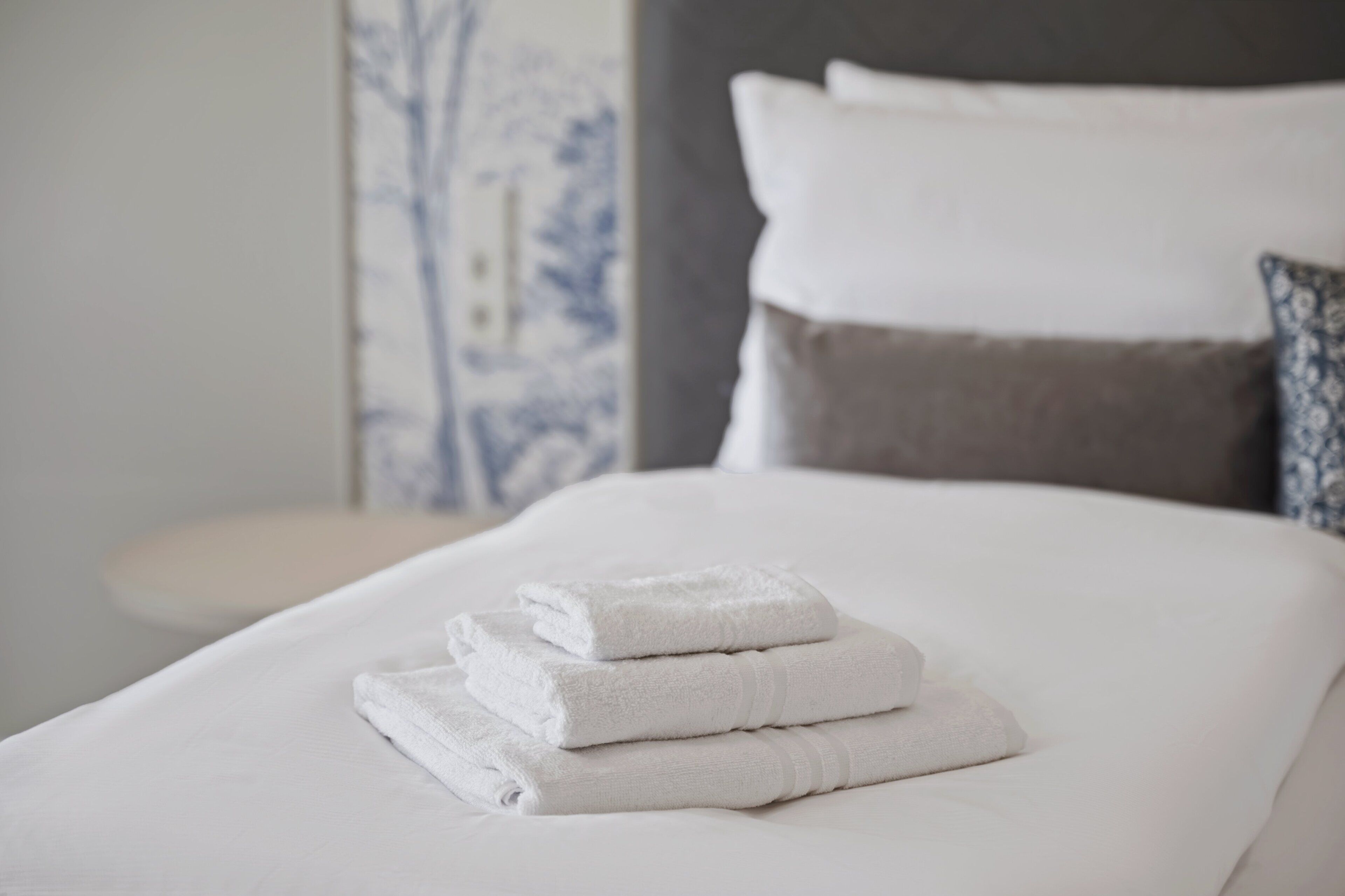 Una cama bien hecha con sábanas blancas y toallas dobladas, una mesita de noche y una pared con un diseño azul y blanco.