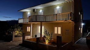 Ein zweistöckiges Haus mit Balkon, Eingangstür und einem kleinen Garten mit Pflanzen.