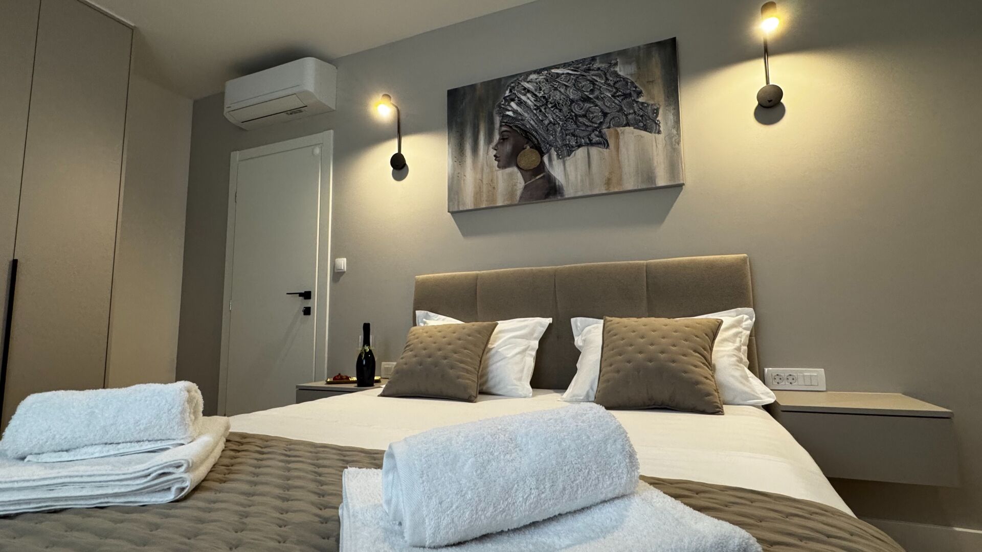 A hotel room with a bed, two bedside tables, a wall-mounted air conditioner, a painting, and a wall-mounted light fixture.