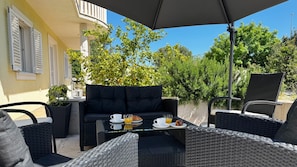 Ein Außenbereich mit Korbmöbeln, einem Tisch mit Frühstücksangeboten und Blick auf Grünflächen und ein Gebäude.