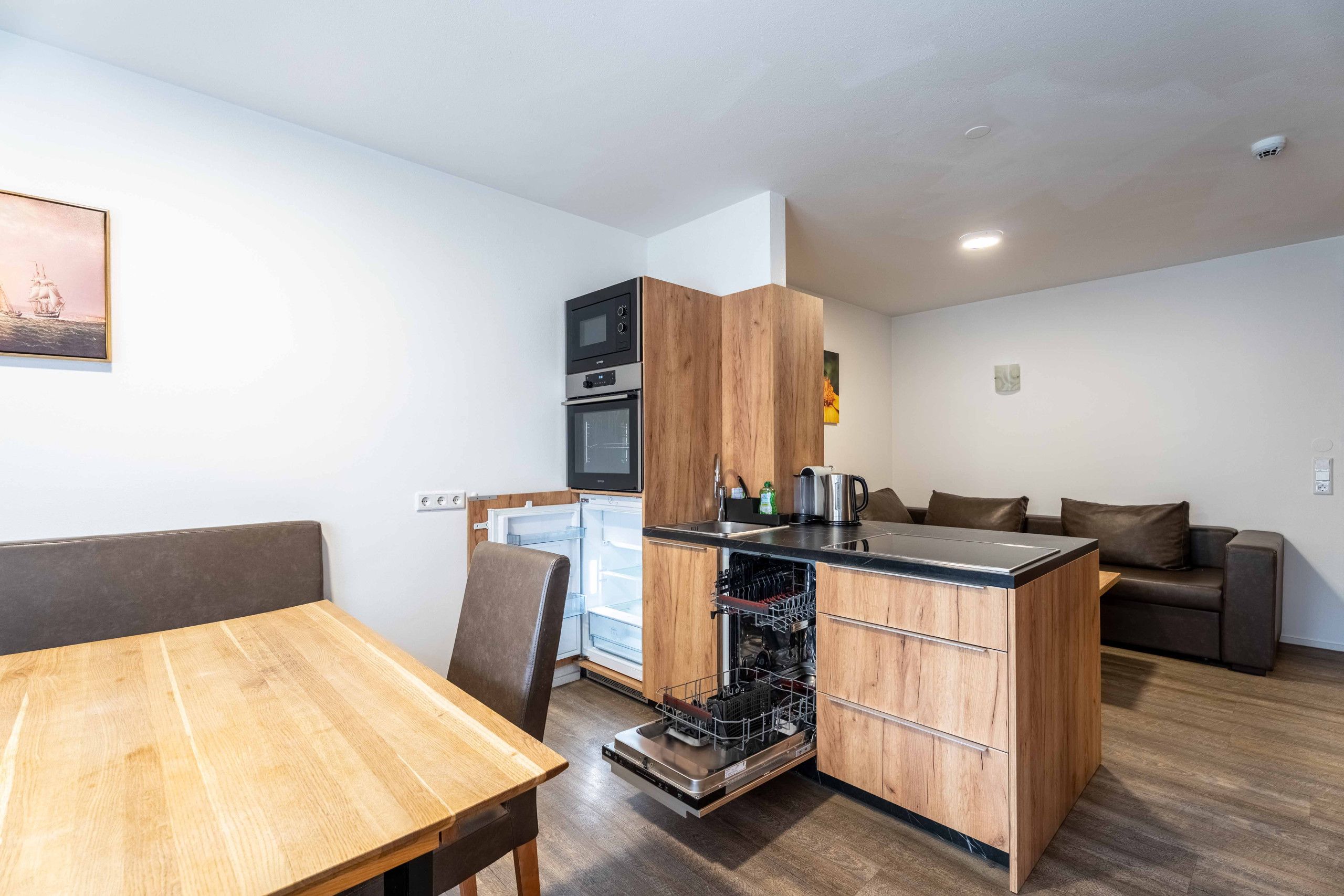 Eine kompakte Küche mit integriertem Backofen, Mikrowelle und Geschirrspüler, einem Holztisch im Essbereich und einer Couch im Wohnbereich.