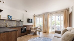 Een moderne keuken met een marmeren aanrechtblad, houten keukenkastjes en een inbouwoven. Er is een eethoek met een ronde tafel en stoelen, en een bank in de woonkamer.