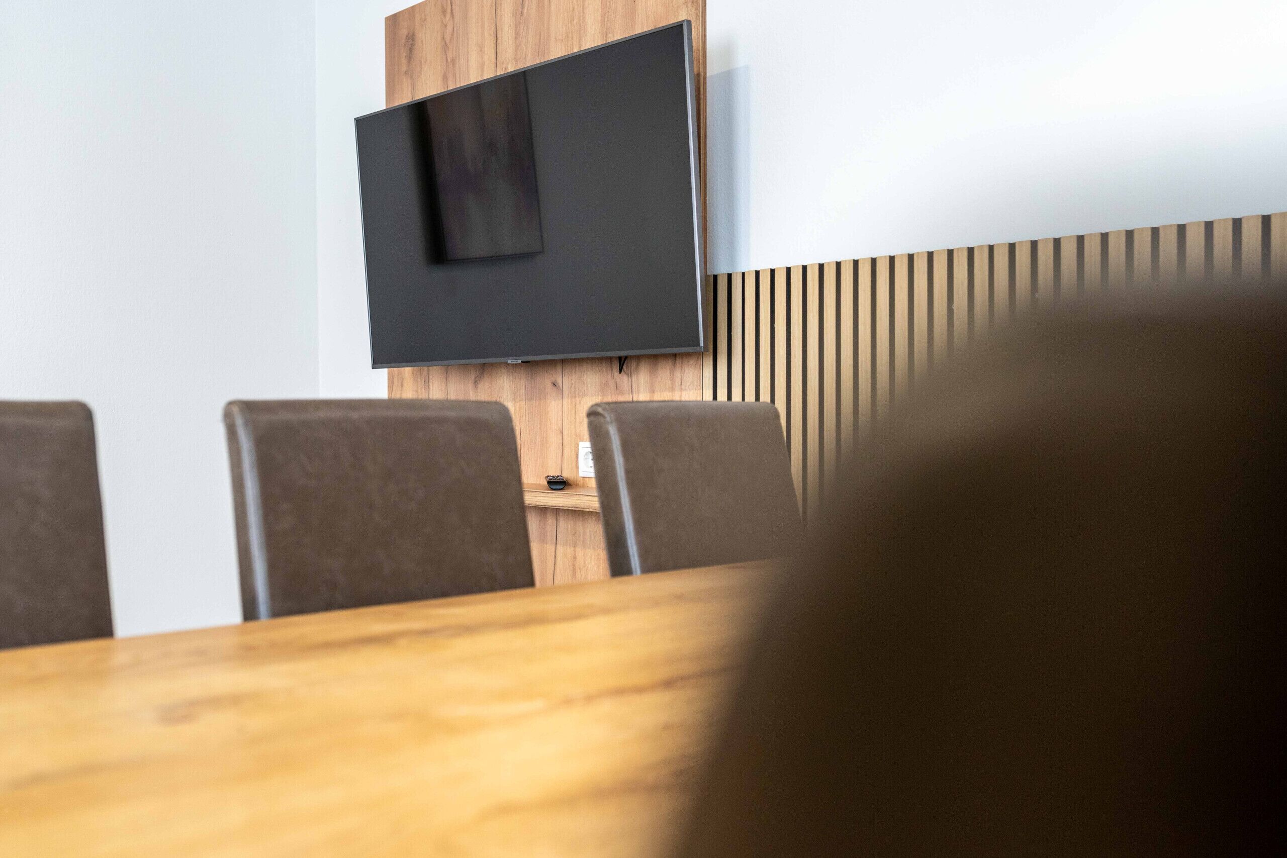 Uma sala de conferências com uma grande televisão de tela plana fixada em uma parede de madeira, uma mesa de madeira e quatro cadeiras.