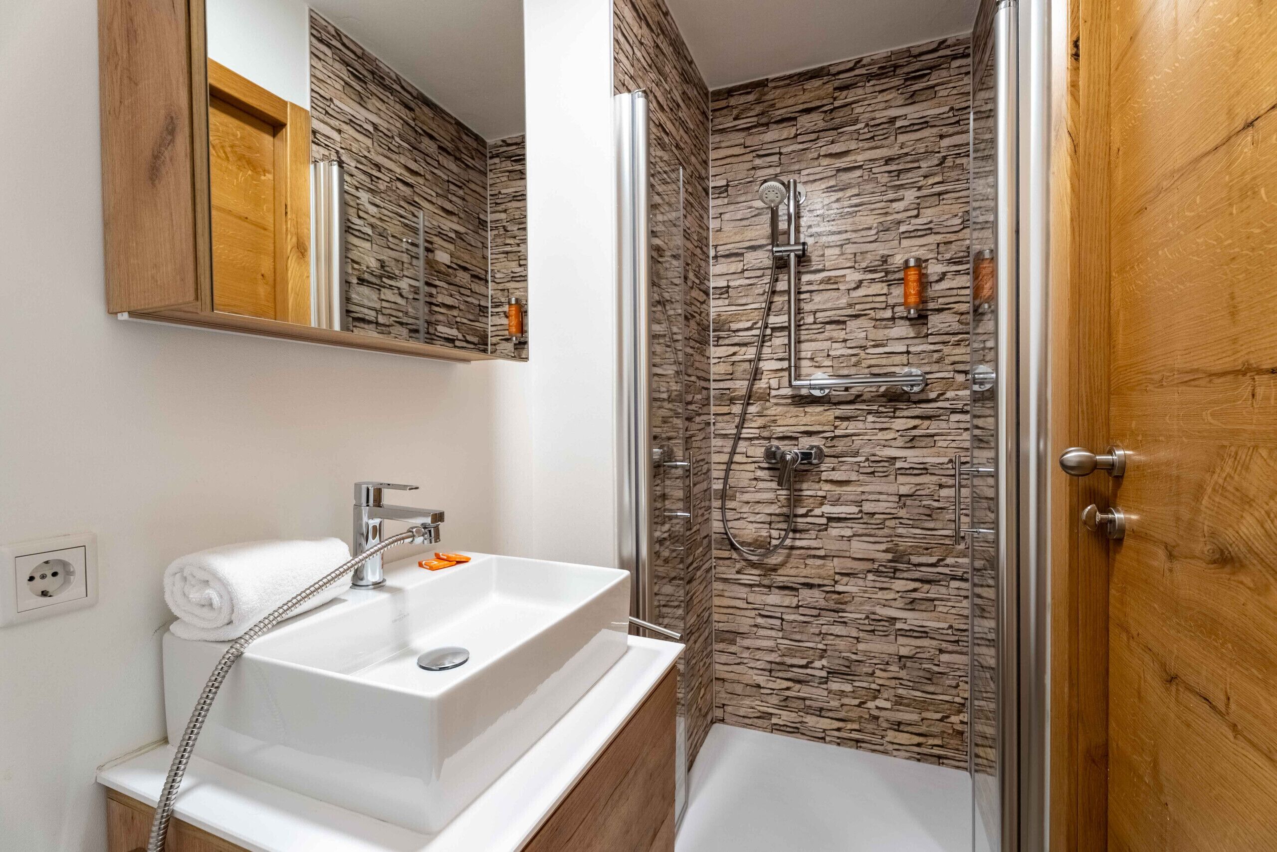 Une salle de bain avec un lavabo blanc, une douche avec un mur en pierre et une porte en bois.