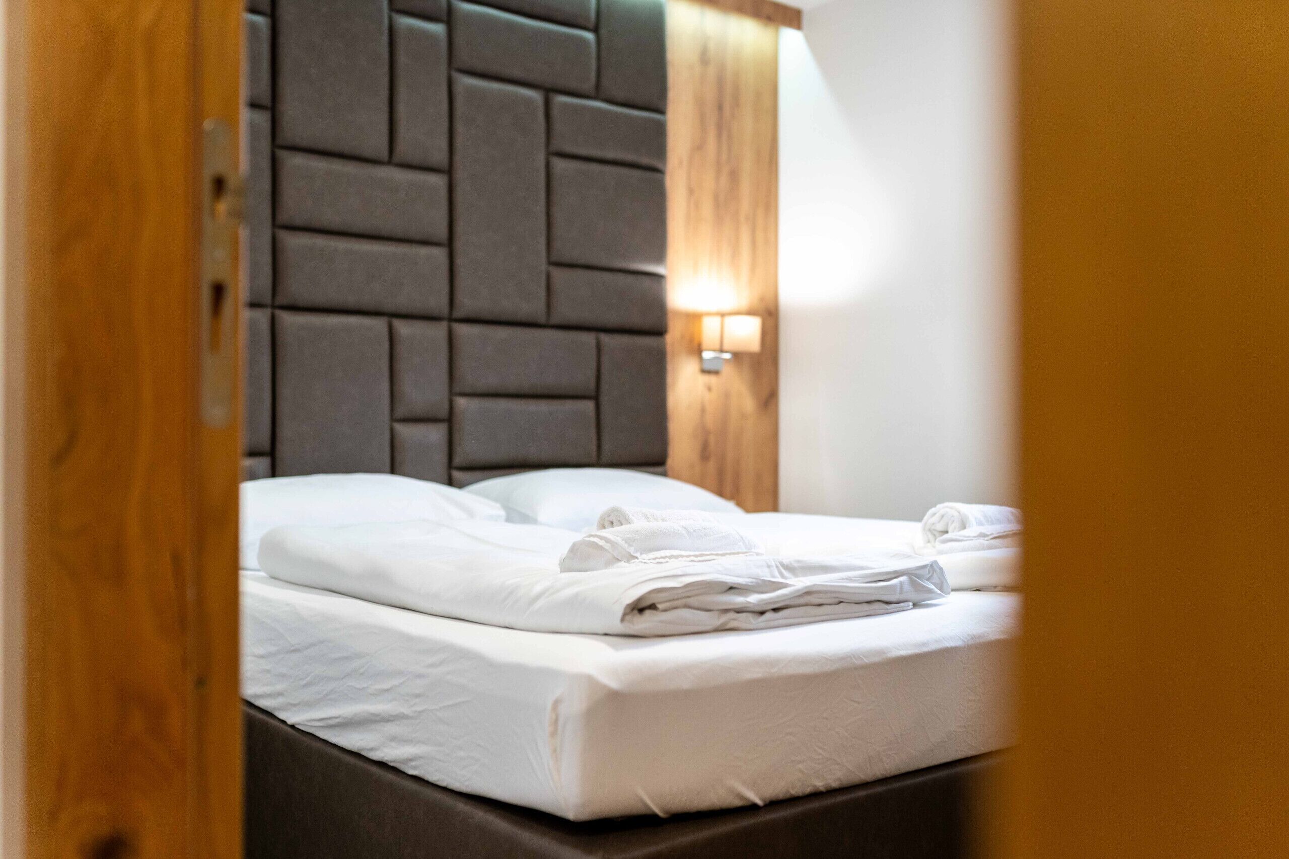 Een net opgemaakt bed met witte lakens, een hoofdbord met een geometrisch patroon en een wandlamp.