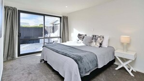 A bedroom with a large sliding glass door, a bed with a grey and white bedspread, a white bedside table with a lamp, and a view of a fenced outdoor area. - Russley home near the airport, single level with parking (Christchurch)