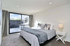Een slaapkamer met een grote schuifdeur, een bed met een grijs-witte dekbedovertrek, een witte nachtkastje met een lamp, en uitzicht op een omheind buitengebied.
